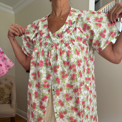 A woman wearing a cream-colored short-sleeve button-front nightgown with a pink and green floral print. She is holding the shoulders of the garment to show its drape, standing in a home interior setting.